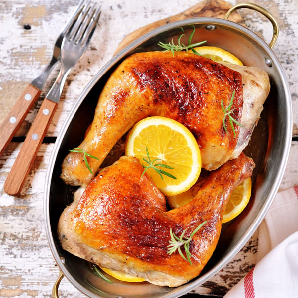 Two golden-brown roasted chicken legs with lemon slices and rosemary in a metal pan.