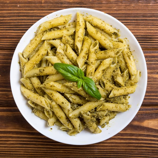 Penne pasta with green pesto sauce and a fresh basil leaf on a white plate.