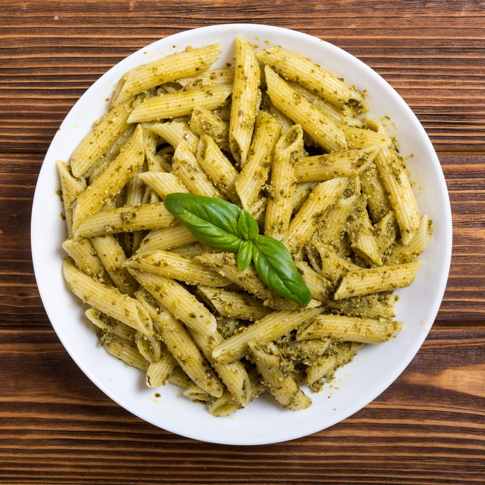 Penne pasta with green pesto sauce and a fresh basil leaf on a white plate.