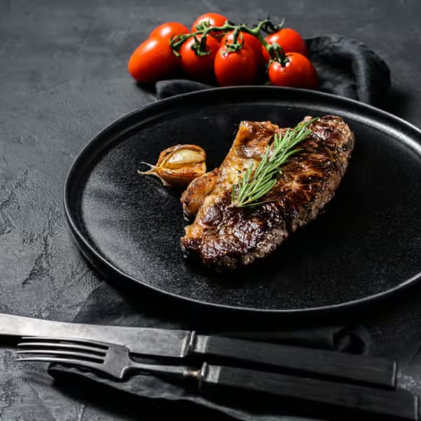Grilled steak with rosemary and garlic on a black plate, with tomatoes and cutlery.
