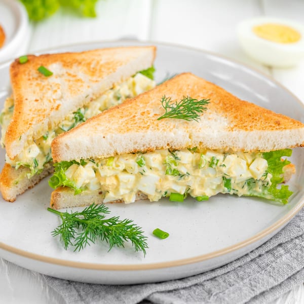 Two toasted egg salad sandwiches with lettuce and dill on a white plate.